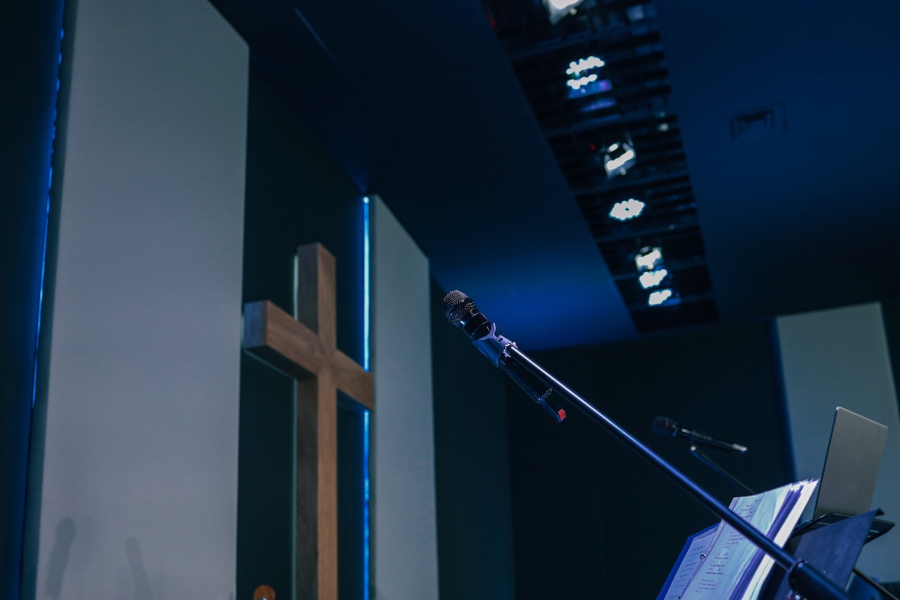 Microphone on a stand with open sheet music and a wooden cross in the background inside a modern worship hall with sleek acoustic panels on the wall with backlighting.